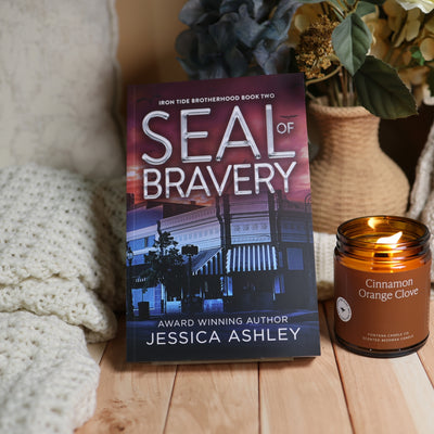 Book cover for 'Seal of Bravery' by Jessica Ashley, styled on a wooden surface next to a lit 'Cinnamon Orange Clove' candle and cozy knitted blankets.