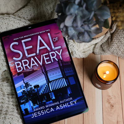 A tablet displaying the book cover of 'Seal of Bravery' by Jessica Ashley, styled on a wooden surface with a knitted blanket, a lit candle, and a potted plant.