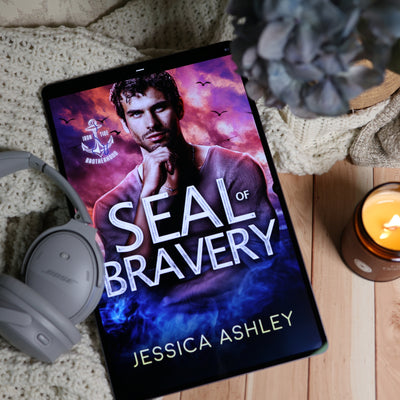 A tablet lying on a wooden surface with a cozy styled setup. The screen displays a book cover titled "SEAL of BRAVERY" by Jessica Ashley, featuring a man's portrait against a dramatic purple and blue sky with birds.