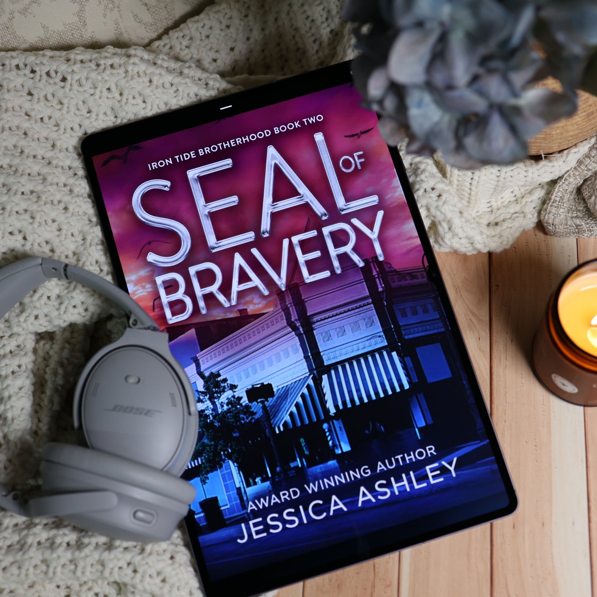 A tablet lying on a knitted blanket with a candle, headphones, and flowers around it, displaying the book cover for 'Seal of Bravery' by Jessica Ashley.