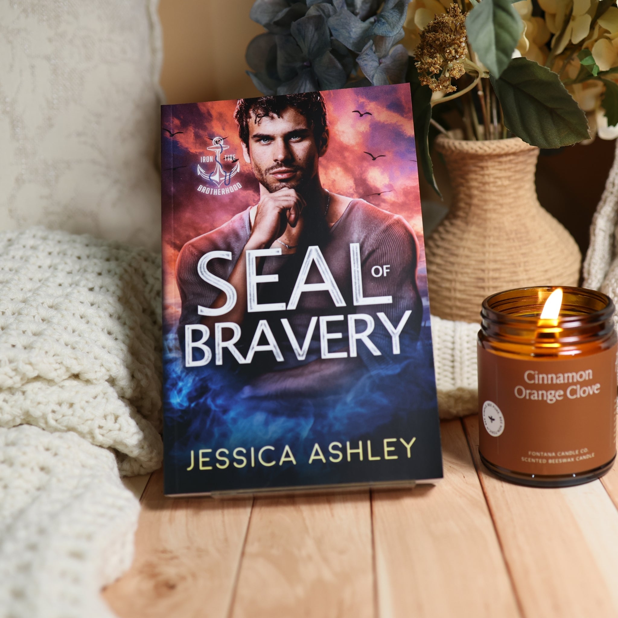 A book titled "Seal of Bravery" by Jessica Ashley stands upright on a wooden surface, styled with a lit candle, knit blanket, and dried flowers in a vase.