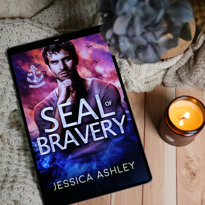 A tablet lying on a wooden surface, displaying the book cover for 'Seal of Bravery' by Jessica Ashley. The scene is styled with a knitted blanket, a potted succulent, and a lit candle.