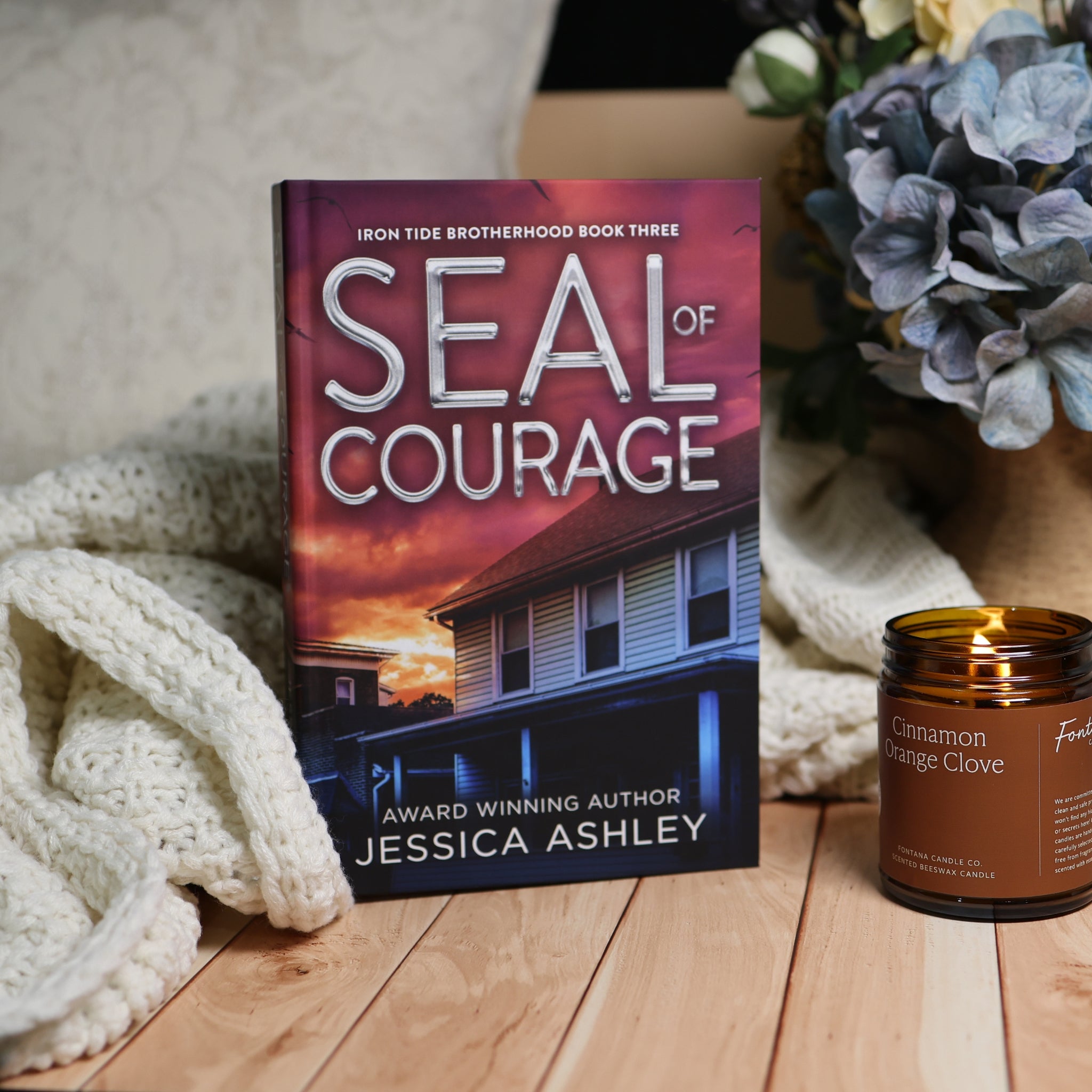 Book cover for 'Seal of Courage' by Jessica Ashley, styled on a wooden surface next to a lit candle and a cream knit blanket, with blue flowers in the background.