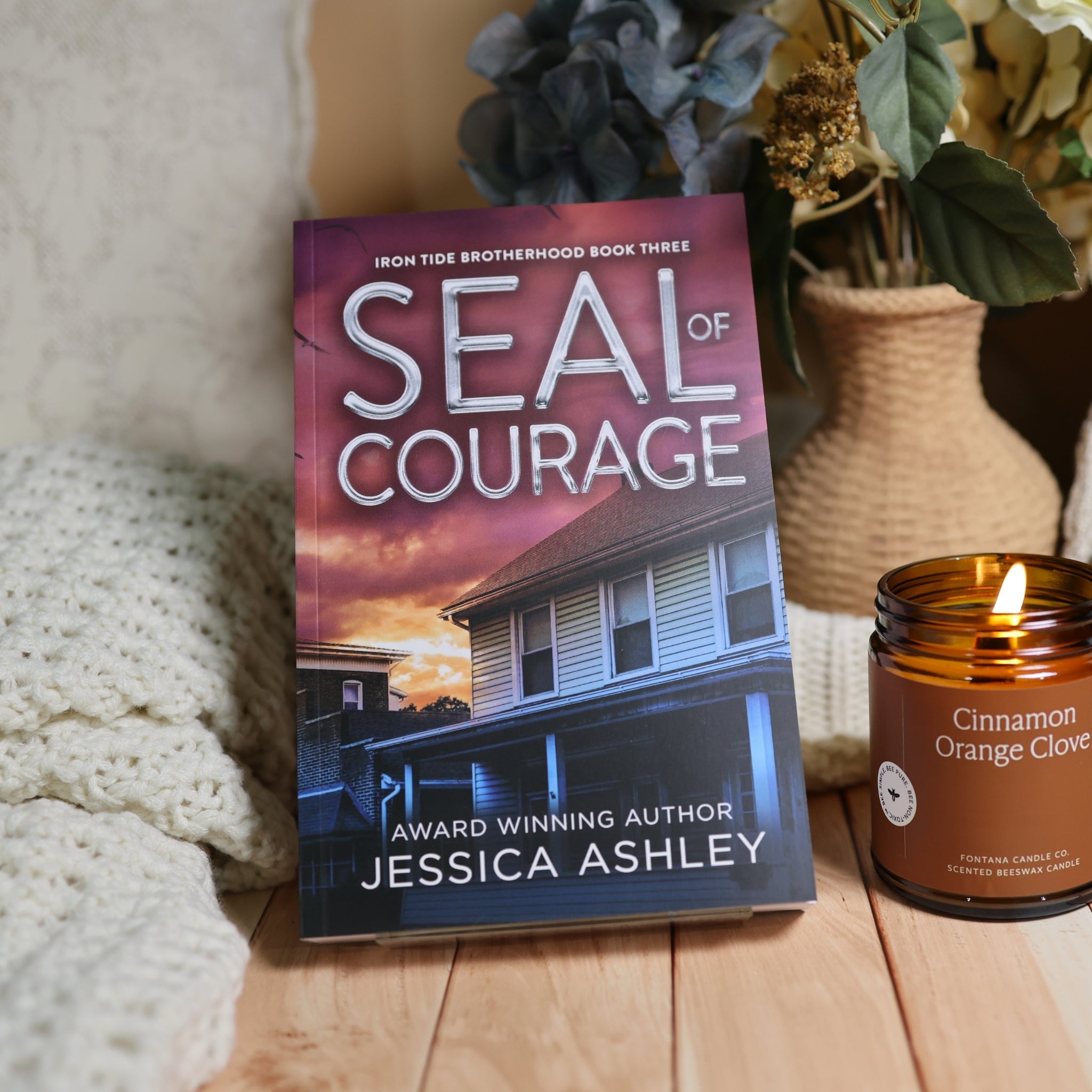 Book cover for 'Seal of Courage' by Jessica Ashley, styled on a wooden surface next to a lit 'Cinnamon Orange Clover' candle and a cozy knit blanket.