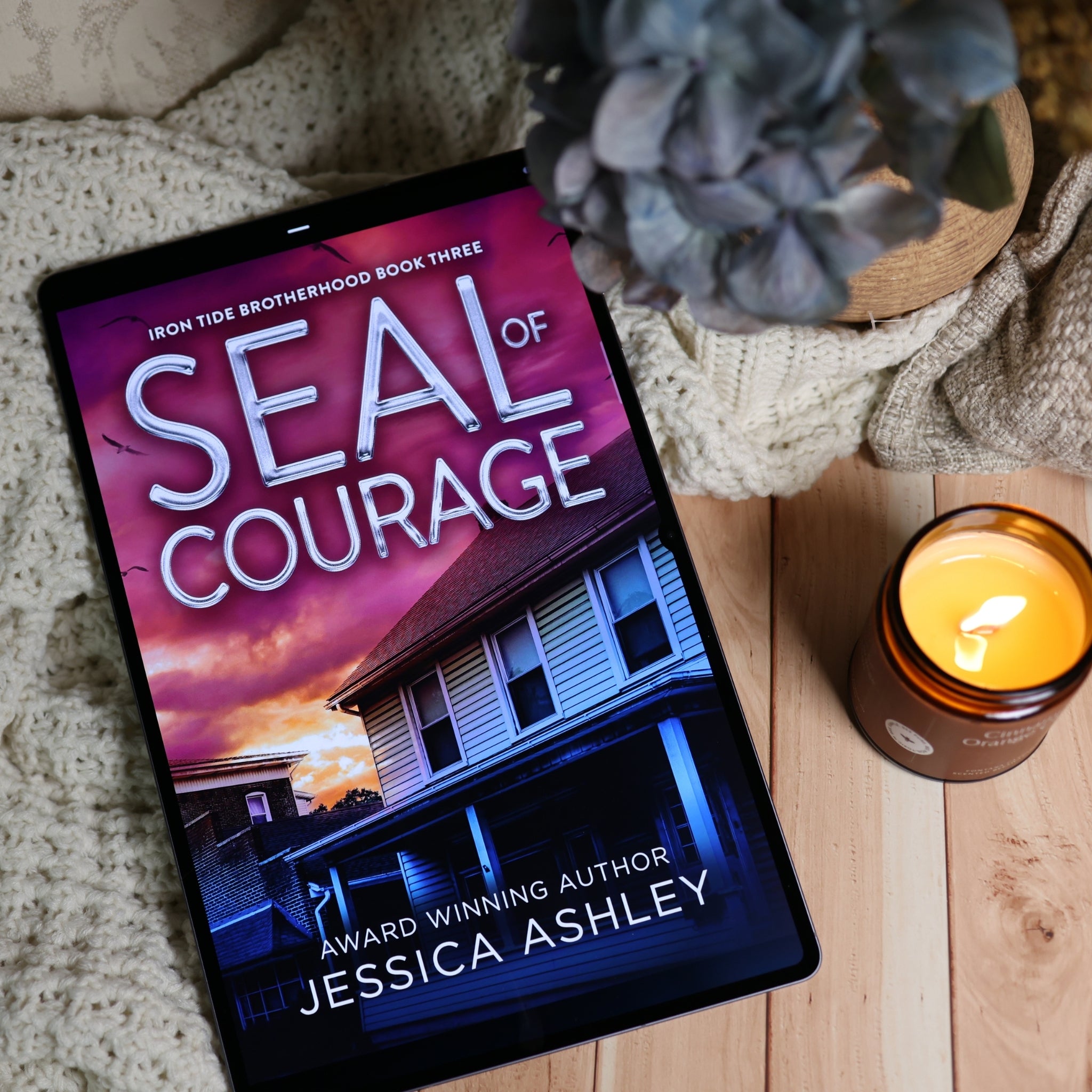 A tablet lying on a wooden surface, displaying the book cover for 'Seal of Courage' by Jessica Ashley. The scene is styled with a knitted blanket, a potted plant, and a lit candle.