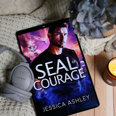A tablet lying on a cream knit blanket on a wooden surface, displaying the book cover for 'Seal of Courage' by Jessica Ashley. A pair of gray Bose headphones rests beside the tablet, with a lit candle and a dried flower nearby.