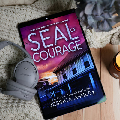 A tablet lying on a cream knit blanket over a wood surface, displaying the book cover for 'Seal of Courage' by Jessica Ashley. Gray Bose headphones rest beside the tablet, with a blurred gray flower and a lit candle nearby.