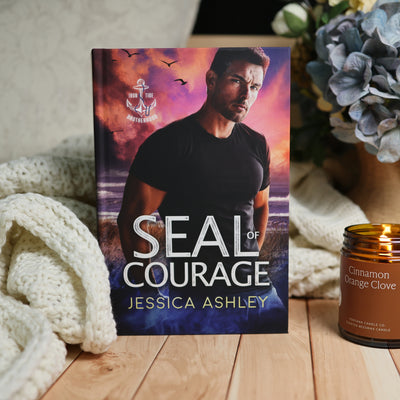 A hardcover book titled "Seal of Courage" by Jessica Ashley stands upright on a wooden surface, styled with a chunky cream knit blanket on the left and a lit amber candle labeled "Cinnamon Orange Clove" on the right. The book cover features a man in a black shirt against a dramatic purple and orange sky with birds and an anchor emblem.