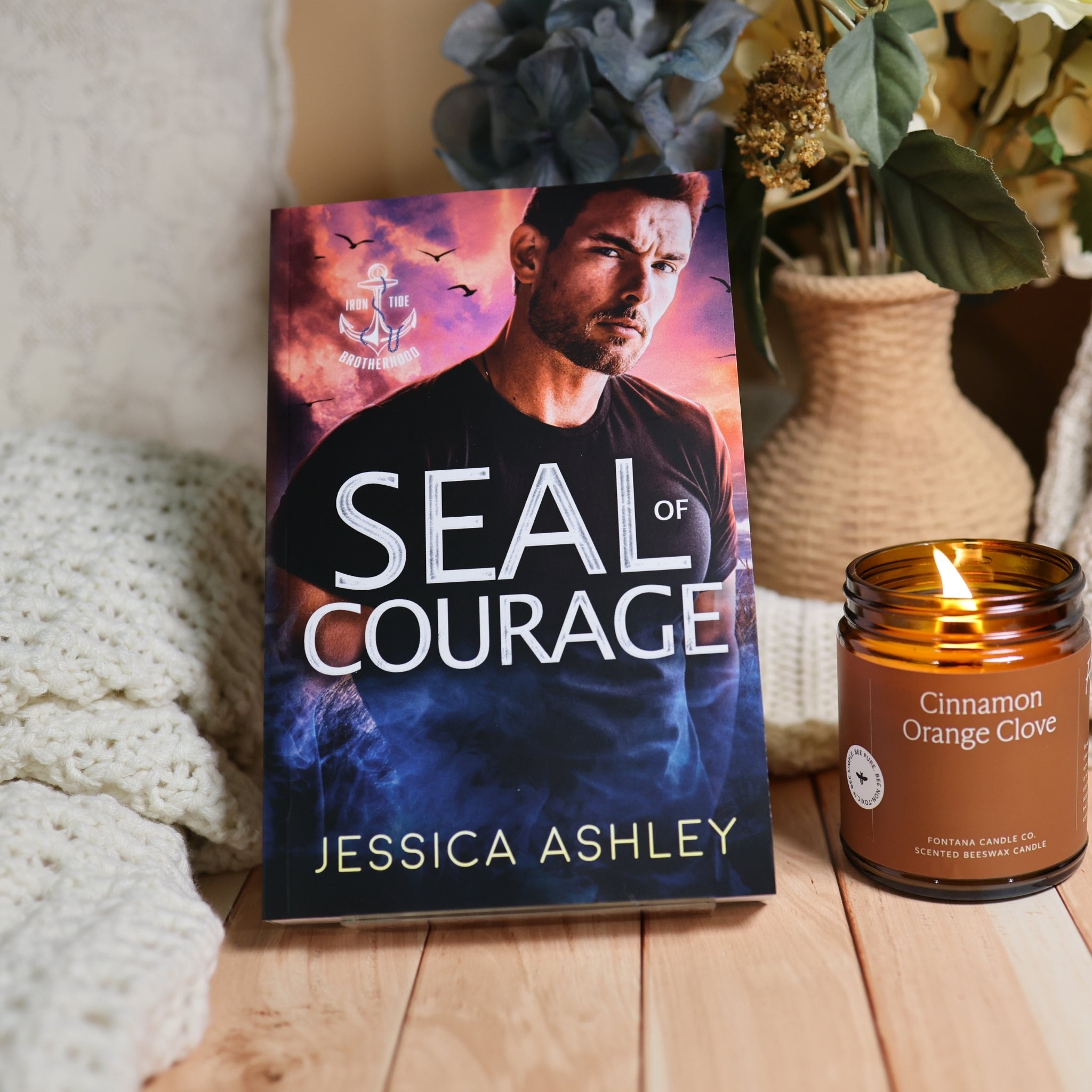 A book titled 'Seal of Courage' by Jessica Ashley stands upright on a wooden surface next to a lit candle in an amber glass jar. The scene includes a knitted cream blanket on the left and a woven vase with dried flowers in the background.