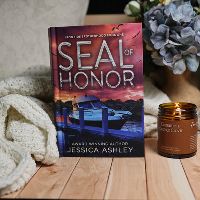 The book 'Seal of Honor' by Jessica Ashley is displayed on a wooden surface next to a lit candle in a glass jar and a cream knit blanket. The book cover features a sunset scene with a boat docked at a pier.