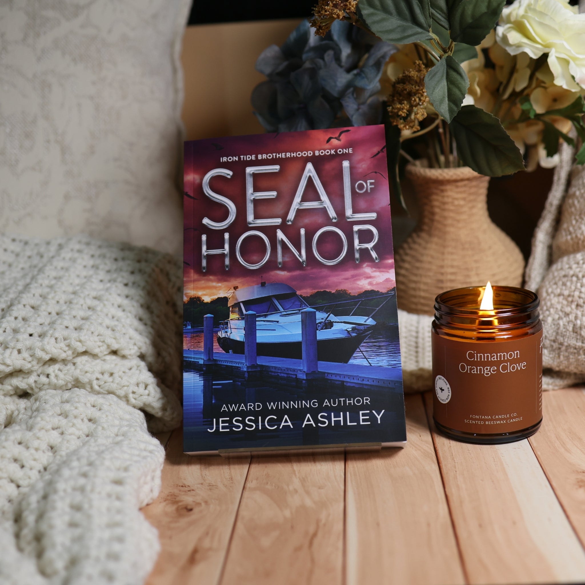 A book titled "Seal of Honor" by Jessica Ashley stands upright on a wooden surface next to a lit candle in a glass jar. The scene includes a knit blanket, dried flowers in a vase, and a warm indoor background.