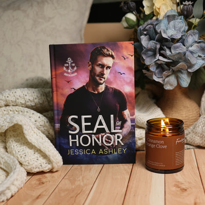 A book titled 'Seal of Honor' by Jessica Ashley stands upright on a wooden surface, styled with a cream knit blanket on the left and a lit amber candle on the right, with dried flowers in the background.