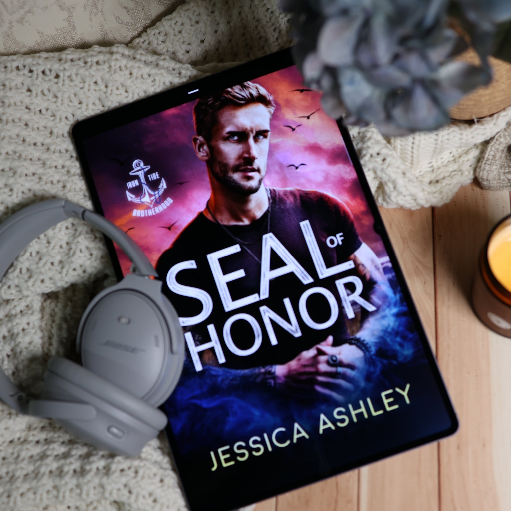 A tablet lying on a knitted blanket and wooden surface, displaying the book cover for 'Seal of Honor' by Jessica Ashley. Gray over-ear headphones rest beside the tablet, with a candle and a blurred plant in the background.