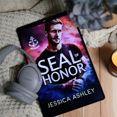 A tablet lying on a knitted blanket and wooden surface, displaying the book cover for 'Seal of Honor' by Jessica Ashley. Gray over-ear headphones rest beside the tablet, with a candle and a blurred plant in the background.
