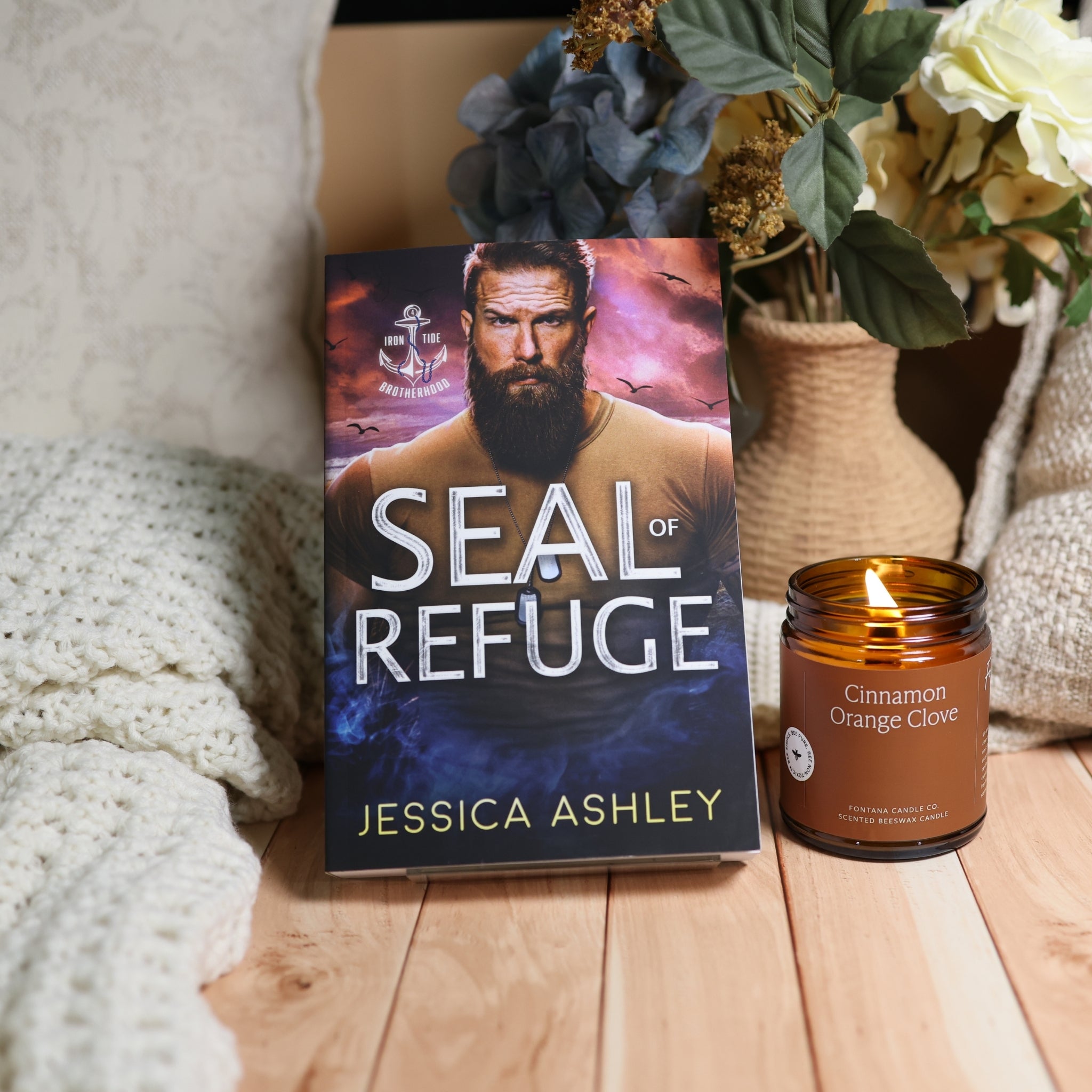 A book titled "SEAL of REFUGE" by Jessica Ashley stands upright on a wooden surface beside a lit amber glass candle labeled "Cinnamon Orange Clove." The scene includes a knitted blanket, dried flowers in a vase, and a cozy indoor setting.