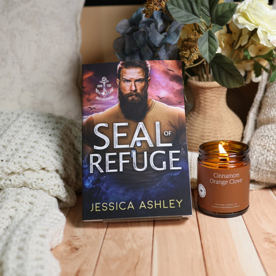 A book titled "SEAL of REFUGE" by Jessica Ashley stands upright on a wooden surface beside a lit amber glass candle labeled "Cinnamon Orange Clove." The scene includes a knitted blanket, dried flowers in a vase, and a cozy indoor setting.