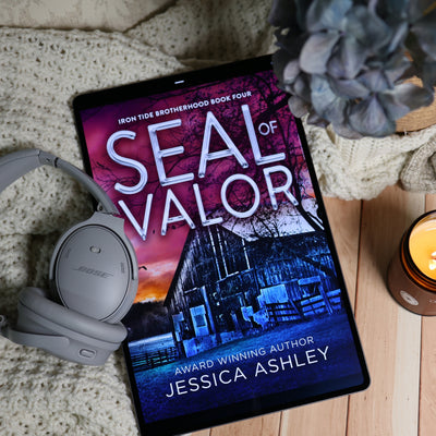A tablet displaying the book cover of 'Seal of Valor' by Jessica Ashley, styled in a cozy flat-lay setting with gray Bose headphones, a knitted blanket, a candle, and a flower on a wooden surface.