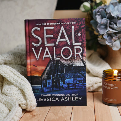 Book cover for 'Seal of Valor' by Jessica Ashley, styled on a wooden surface with a cream knit blanket, a lit candle, and blue flowers in the background.