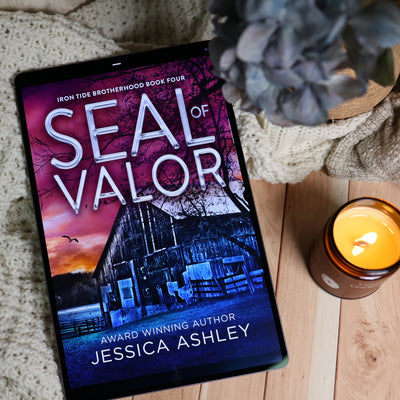 A tablet displaying the book cover of 'Seal of Valor' by Jessica Ashley, styled on a wooden surface with a knitted blanket, a lit candle, and a blue hydrangea flower.