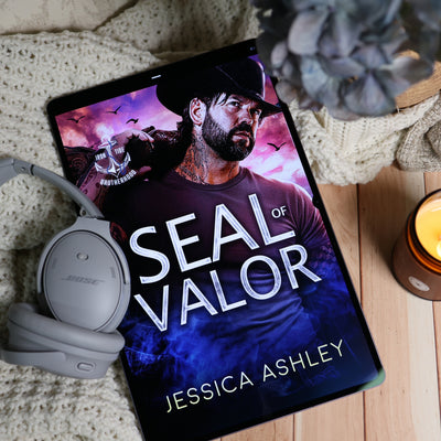 A tablet lying on a knitted blanket and wood surface, displaying a book cover titled "SEAL OF VALOR" by Jessica Ashley. White over-ear headphones rest beside the tablet, with a gray flower and a lit candle nearby.