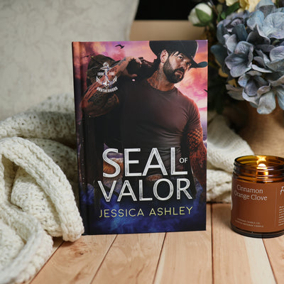 A book titled "Seal of Valor" by Jessica Ashley stands upright on a wooden surface, styled with a cream knit blanket on the left and a lit amber candle on the right. The cover shows a bearded man in a black cap against a pink and blue sky background.