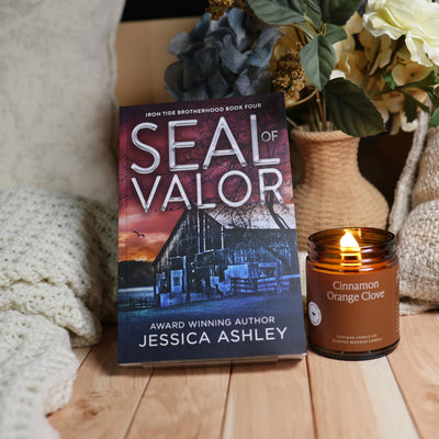 A book titled "Seal of Valor" by Jessica Ashley stands upright on a wooden surface next to a lit amber glass candle labeled "Cinnamon Orange Clove." The scene includes a cozy knit blanket, flowers in a vase, and a neutral indoor background.