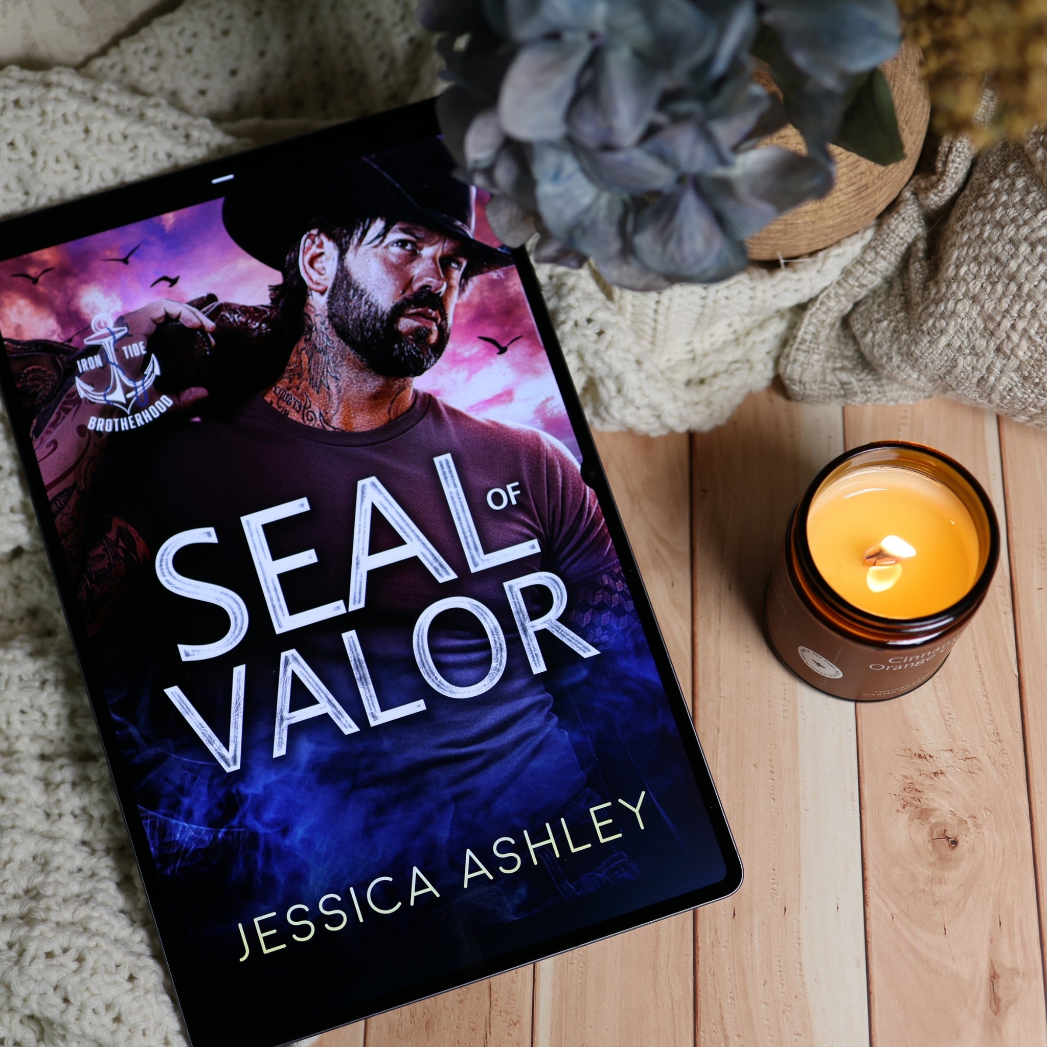 A tablet lying on a light wood surface, displaying the book cover for 'Seal of Valor' by Jessica Ashley. The scene is styled with a knitted blanket, a lit amber candle, and dried flowers in the background.