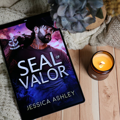 A tablet lying on a light wood surface, displaying the book cover for 'Seal of Valor' by Jessica Ashley. The scene is styled with a knitted blanket, a lit amber candle, and dried flowers in the background.