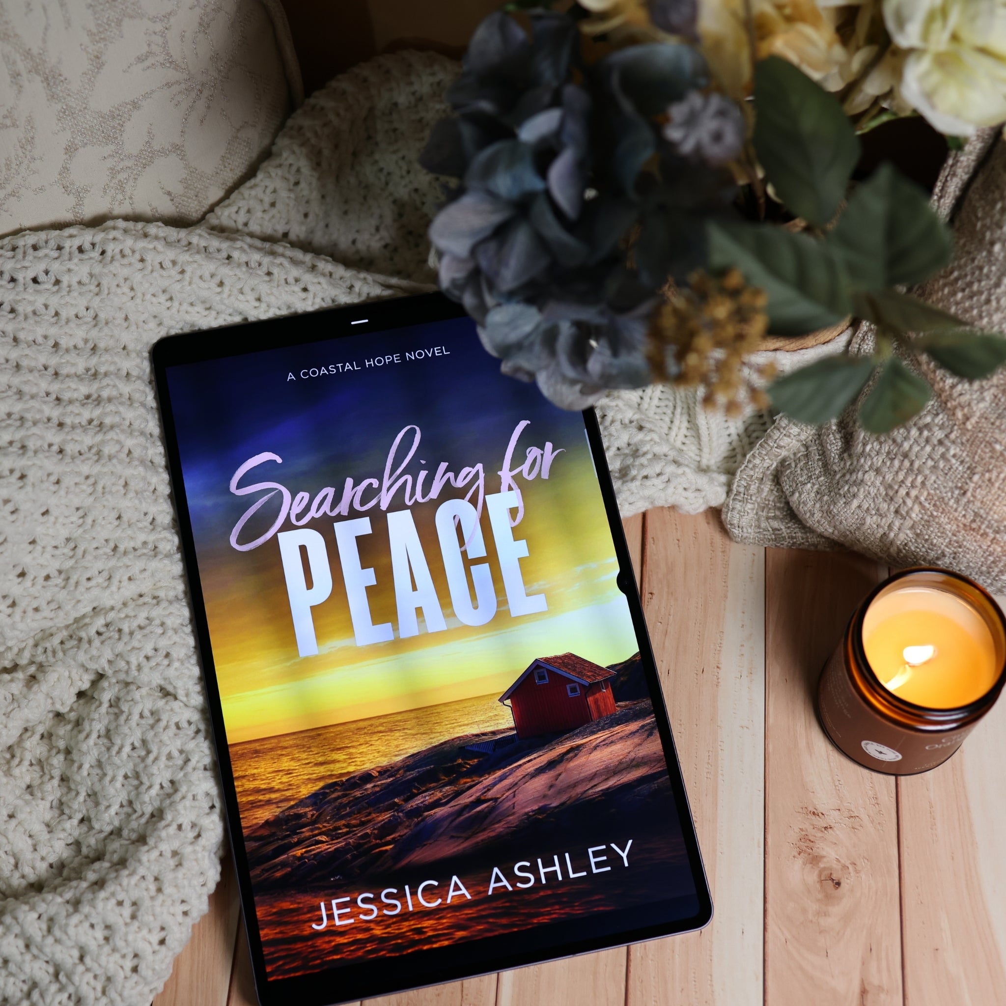 A tablet lying on a wooden surface, displaying the book cover for 'Searching for Peace' by Jessica Ashley. The scene is styled with a cream knit blanket, a lit amber candle, and a bouquet of blue and cream flowers.