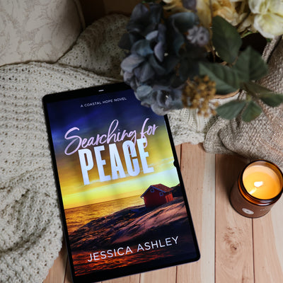 A tablet lying on a wooden surface, displaying the book cover for 'Searching for Peace' by Jessica Ashley. The scene is styled with a cream knit blanket, a lit amber candle, and a bouquet of blue and cream flowers.