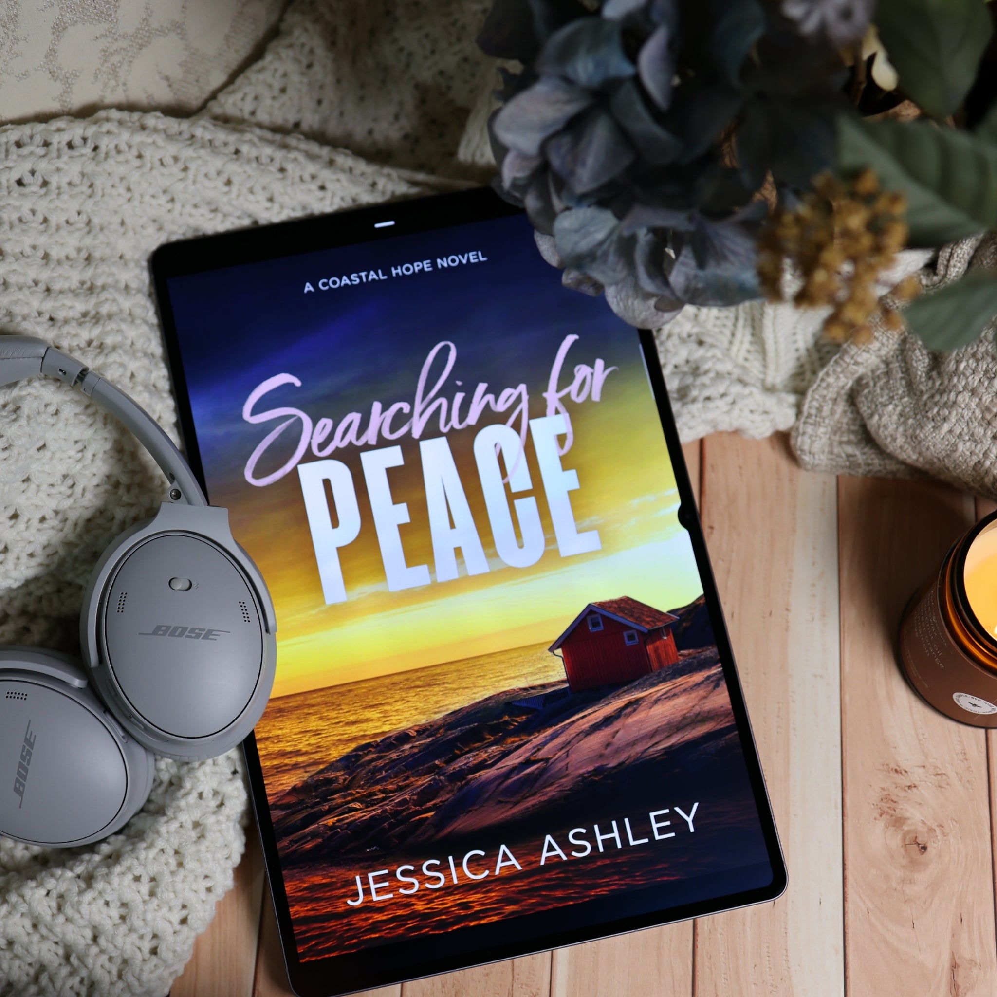 A tablet lying on a wooden surface, displaying the book cover for "Searching for Peace" by Jessica Ashley. The scene is styled with a knit blanket, gray Bose headphones, dried flowers, and a candle, creating a cozy reading atmosphere.