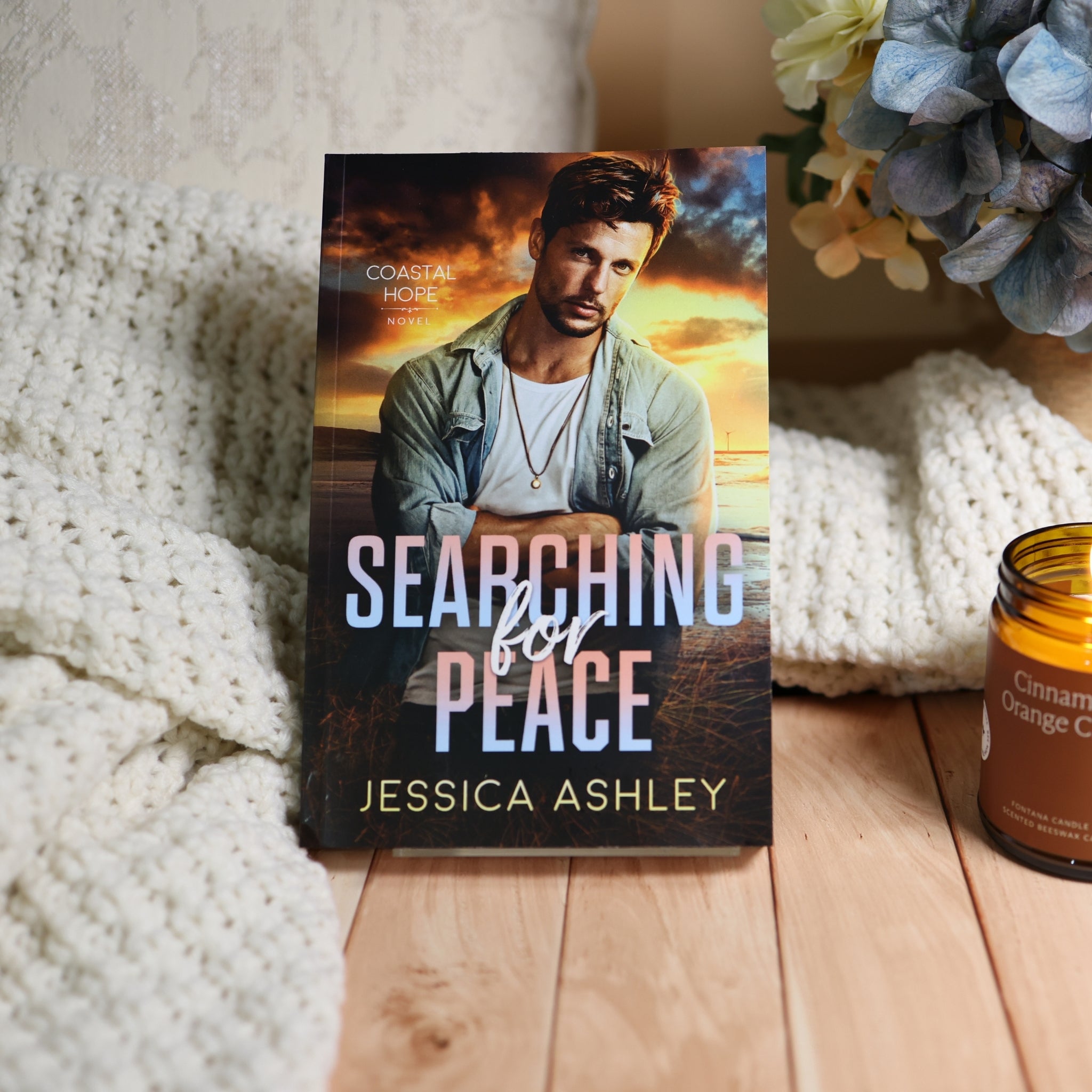 A romance novel book titled 'Searching for Peace' by Jessica Ashley, standing upright on a light wood surface next to a white knit blanket, flowers, and an amber candle jar.