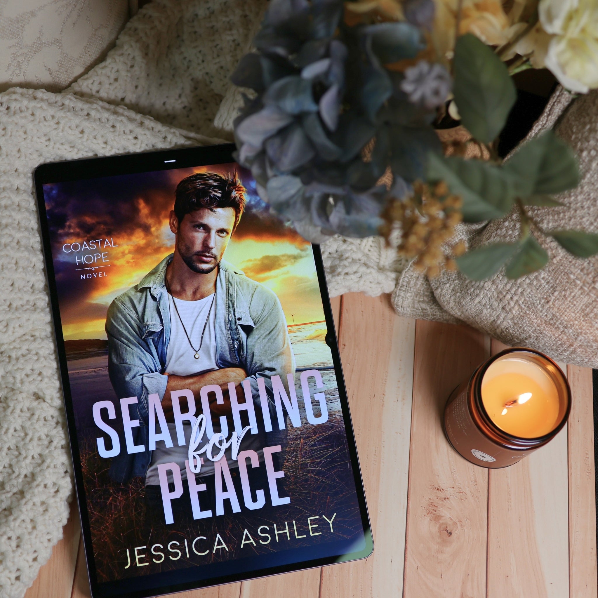 A tablet displaying the book cover of 'Searching for Peace' by Jessica Ashley, styled on a wooden surface with a lit candle and flowers.