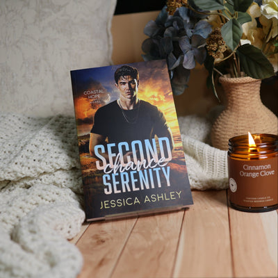A book titled "Second Chance Serenity" by Jessica Ashley is displayed upright on a wooden surface, styled with a cream knit blanket, a lit candle, and dried flowers in the background.