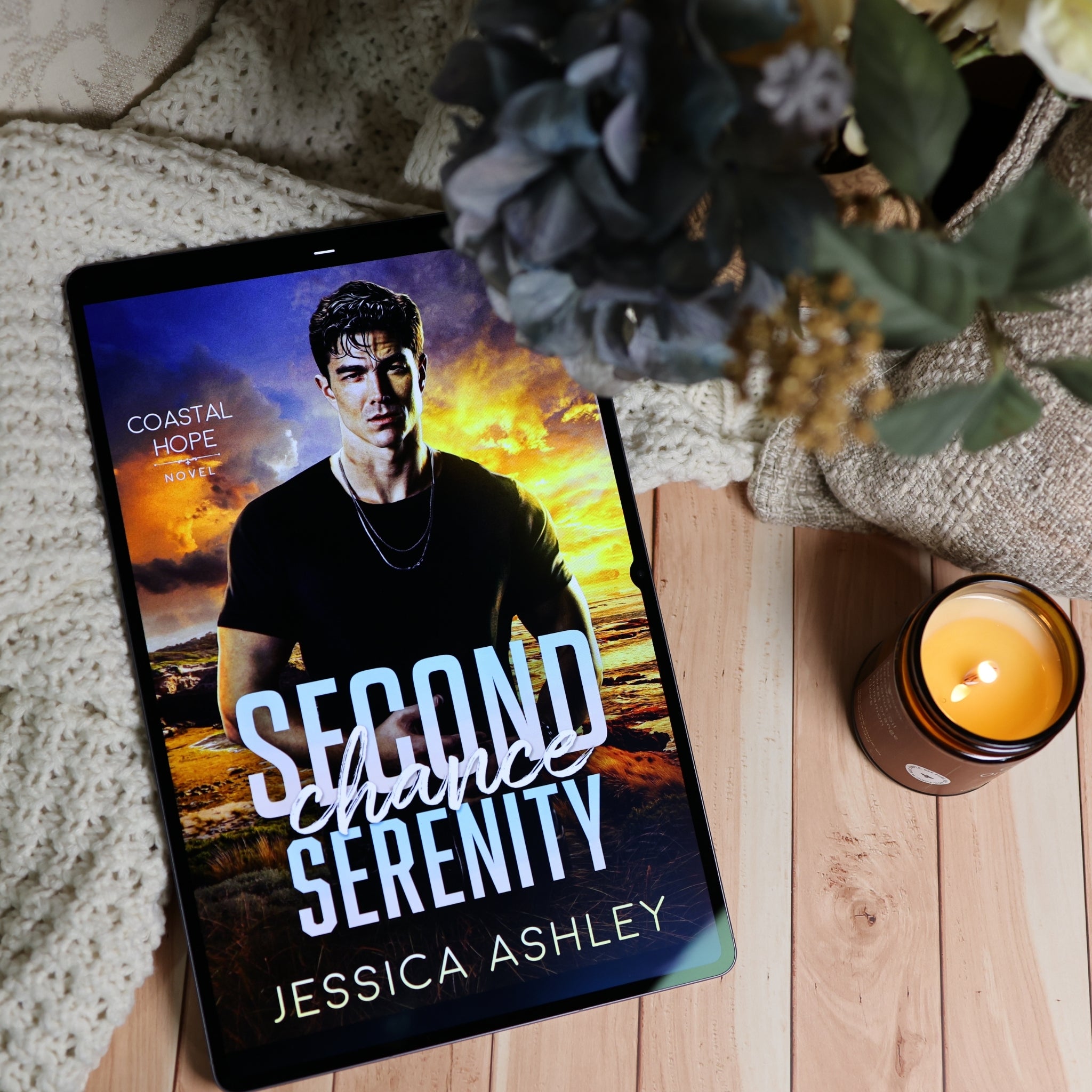A tablet lying on a wooden surface, displaying a romance novel cover titled "Second Chance Serenity" by Jessica Ashley. The scene is styled with a textured cream knit blanket, a lit amber candle, and dried flowers in a vase.
