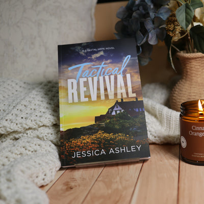 A book titled "Tactical Revival" by Jessica Ashley stands upright on a wooden surface next to a lit amber candle and a cream knit blanket, with dried flowers in the background.