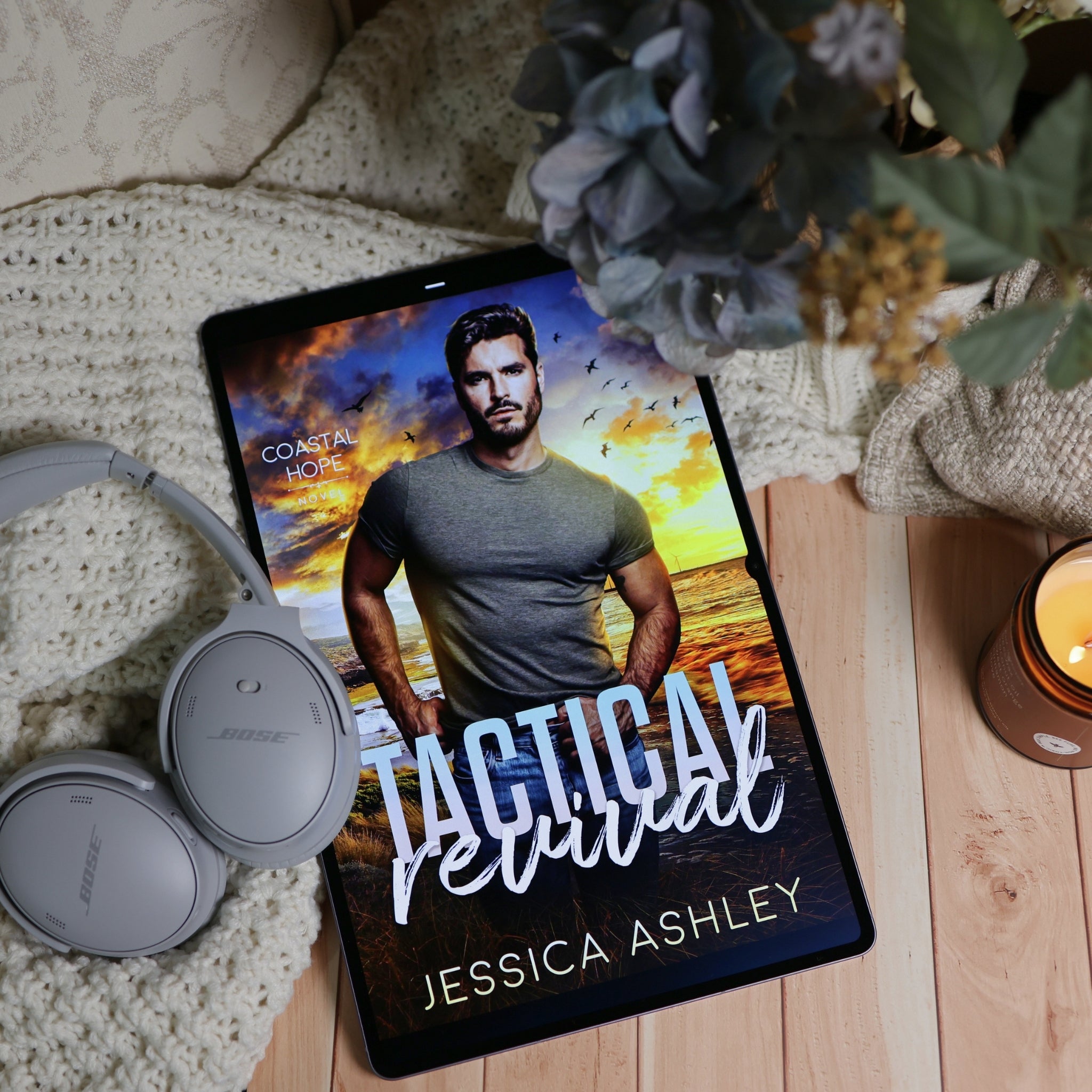 A tablet lying on a wooden surface, displaying a romance book cover titled "Tactical Revival" by Jessica Ashley. The scene is styled with a cream knit blanket, gray over-ear headphones, a lit candle, and dried flowers around the tablet.