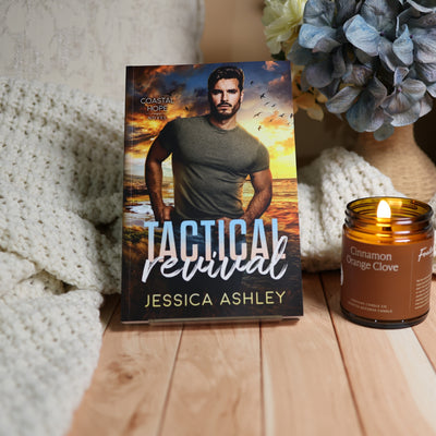 A romance novel book titled 'Tactical Revelation' by Jessica Ashley stands upright on a wooden surface, styled with a knitted cream blanket, a lit candle, and blue flowers in the background.