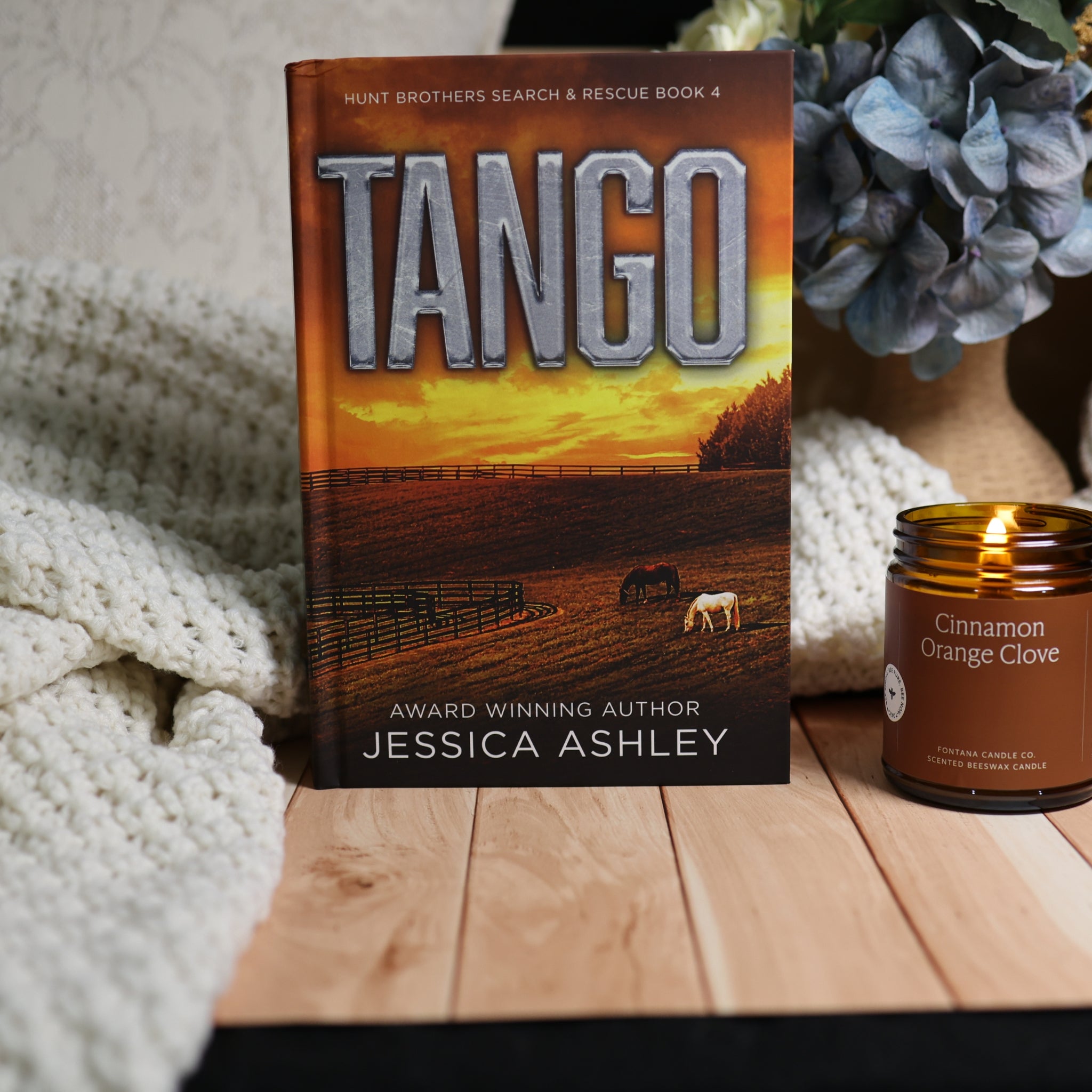The book 'Tango' by Jessica Ashley stands upright on a wooden surface, styled with a cozy white knit blanket on the left and a lit 'Cinnamon Orange Glove' candle on the right. The book cover features a sunset landscape with horses.