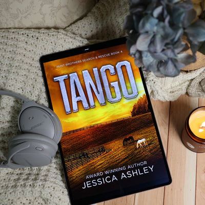 A tablet lying on a beige knit blanket, displaying the book cover for 'Tango' by Jessica Ashley. The scene is styled with gray Bose headphones, a blue-gray hydrangea, and a lit candle on a light wood surface.