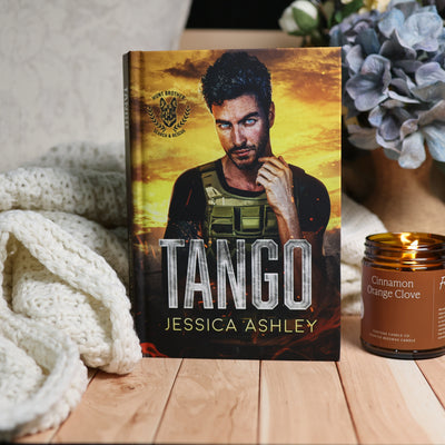 A hardcover book titled 'Tango' by Jessica Ashley stands upright on a wooden surface, styled with a white knit blanket on the left and a lit candle on the right. The book cover features a man in tactical gear against a fiery yellow and orange sky.