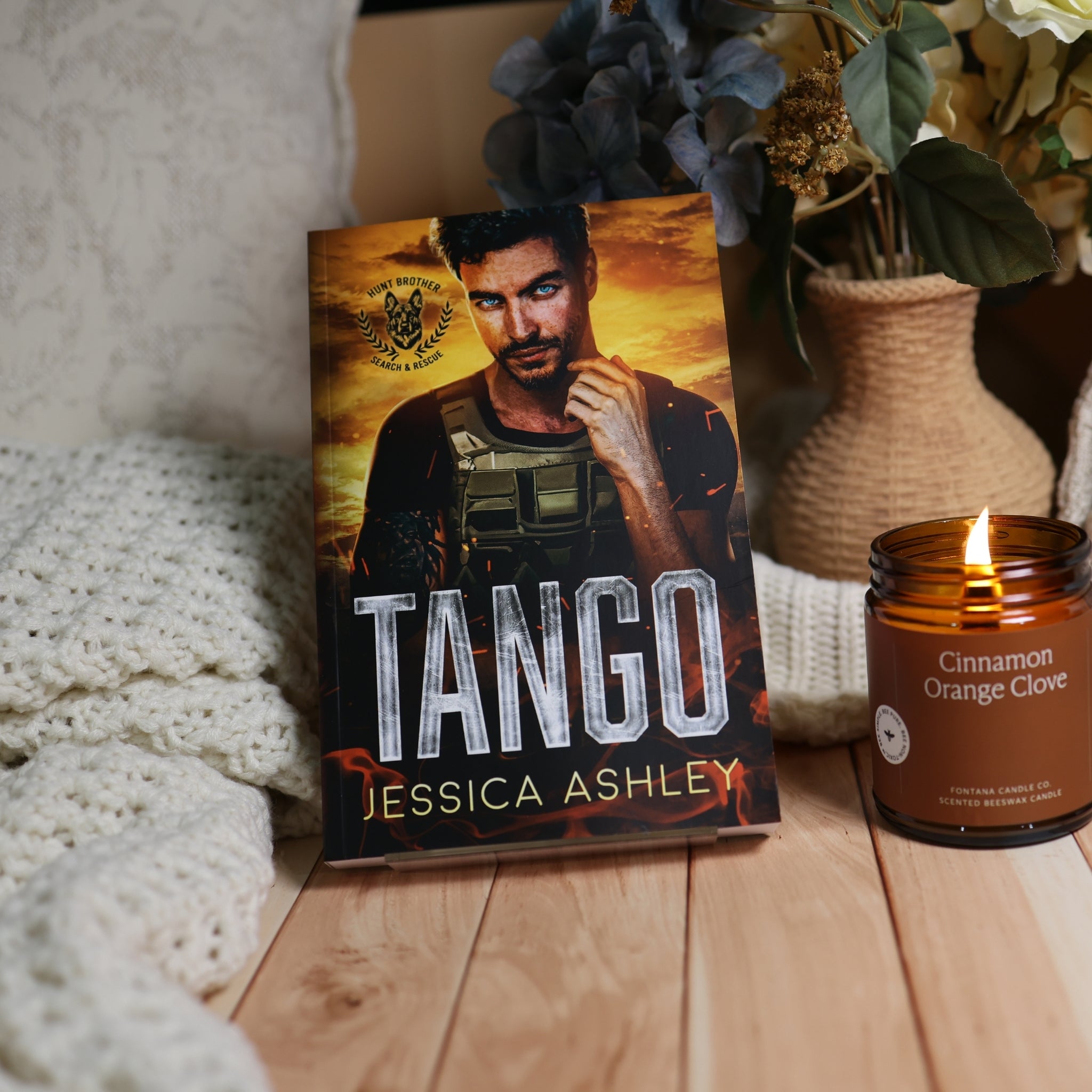 A hardcover book titled 'TANGO' by Jessica Ashley stands upright on a wooden surface, styled with a white knit blanket and a lit candle in the background.